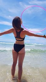 Rear view of woman with arms outstretched playing with hula hoop while standing in sea against sky