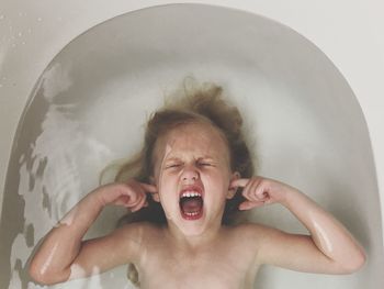 Directly above shot of shouting shirtless girl covering her ears in bathtub