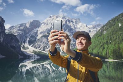 Smiling hiker taking selfie through mobile phone against lake and mountains