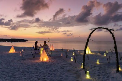 Couple sitting amidst illuminated lighting equipment at beach against sky during sunset