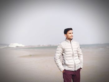 Young man looking away while standing at beach against clear sky