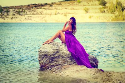 Full length of beautiful woman in purple dress sitting on rock at lakeshore