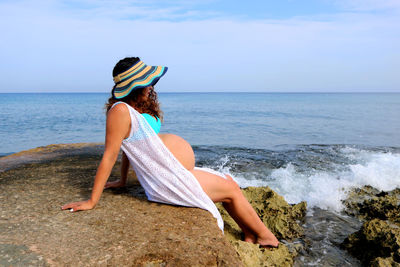 Pregnant woman sitting on rock at beach against sky