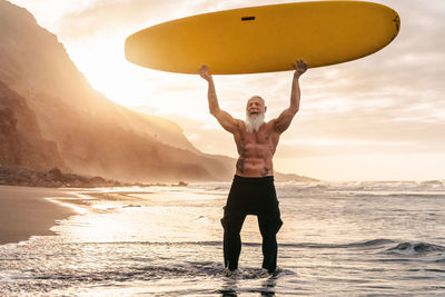 Full length of shirtless man lifting surfboard while standing in sea