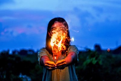 Low angle view of young woman holding sparklers while standing against blue sky at dusk