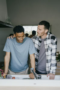 Man standing with hand on friend's shoulder in kitchen seen through window