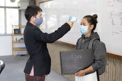 Side view of man wearing flu mask checking temperature of woman at school