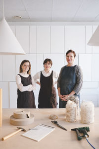 Portrait of confident design professionals wearing aprons with various equipment on desk in workshop