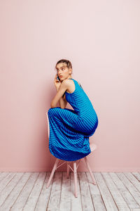 Side view of young woman crouched on top of a chair looking away while talking on the mobile phone on pink background