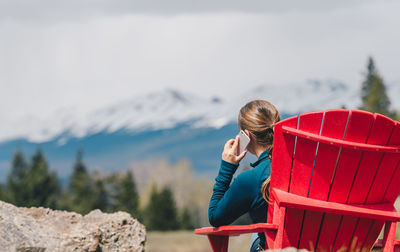Rear view of woman using smart phone while sitting on chair against snowcapped mountain