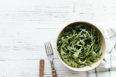 Green fresh arugula salad leaves in bowl, fork and knife on white wooden background, white towel