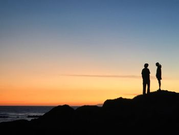 Silhouette friends standing on rock against sea during sunset