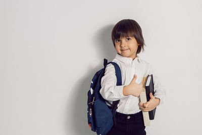 Child boy with a book textbook and backpack stands on a white background thumbs up