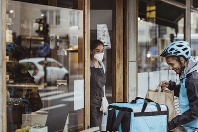 Smiling delivery man collecting order from female owner at delicatessen shop during pandemic