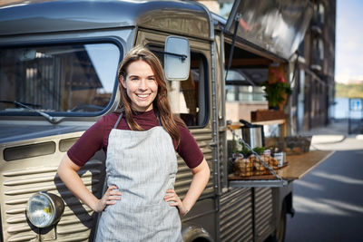 Portrait of smiling female owner standing hands on hips outside food truck in city