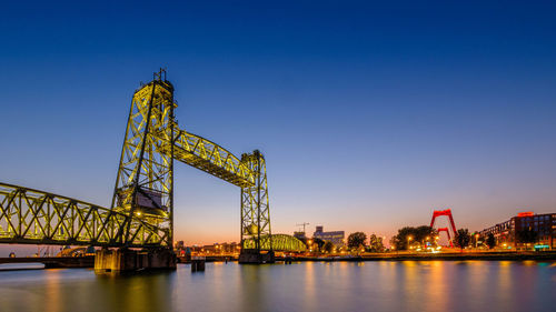 Illuminated cranes at riverbank against clear blue sky