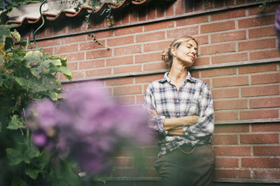 Smiling mature woman with eyes closed and arms crossed leaning on wall