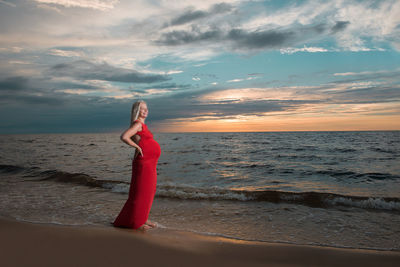 Side view of pregnant woman standing at beach against cloudy sky during sunset