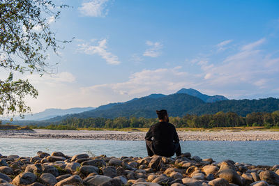 Rear view of man looking at mountains while sitting on rock by river