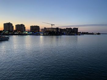 Sea by illuminated buildings against clear sky during sunset