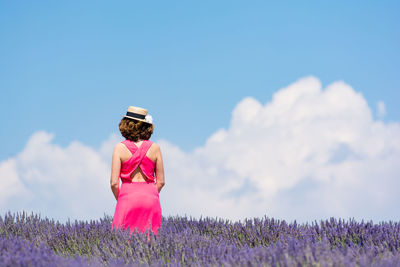 Woman in pink dress with hat standing in lavender field against sky during sunny day