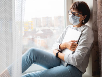 Woman in medical mask had lost her job. she sits on window sill with closed laptop. 