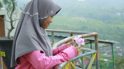 Side view of teenage girl wearing hijab holding pink flowers by railing