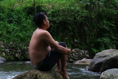 Side view of shirtless man sitting on rock in forest