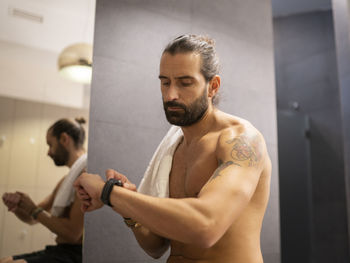 Adult shirtless sportsman with towel checking pulse on smart watch near mirror in locker room in gym