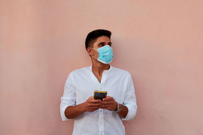 Young latin man with mask is looking his phone on pink background