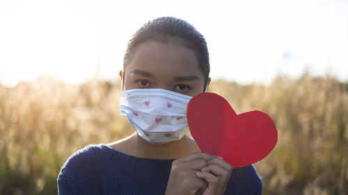 Portrait of girl wearing mask holding heart shape while standing outdoors