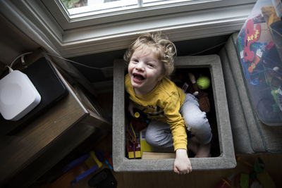 Laughing boy sits in toy box next to storage bins looks up at camera
