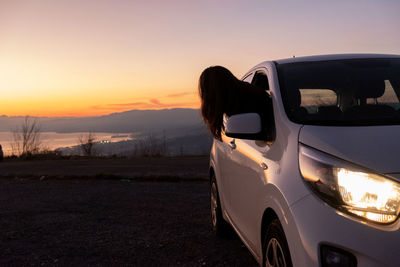 Woman standing by car on field against sky during sunset