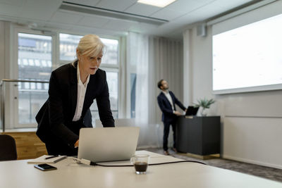 Businesswoman using laptop while colleague standing by flat screen during global conference meeting in office