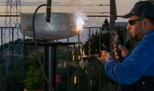 Mid adult man welding metal by railing during sunset