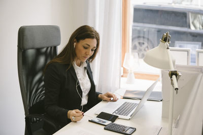 Mature female realtor listening to headphones while looking at laptop in office