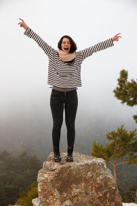 Full length of carefree young woman with arms raised standing on rock during foggy weather