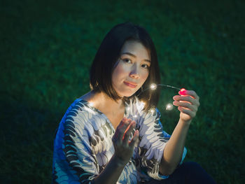 Young woman holding illuminated string lights while sitting on grassy field