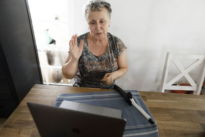 Senior woman sitting at table at home with laptop and curling iron