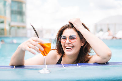 Stylish young woman plus size in black swimsuit and sunglasses with glass of cocktail in pool