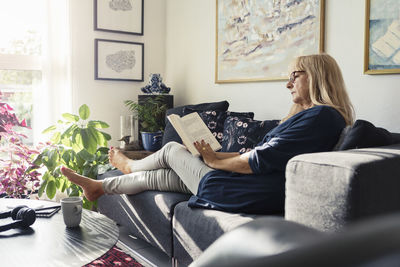 Blond woman reading book while sitting on sofa in living room