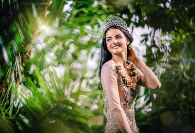 Smiling beautiful bride looking away while wearing crown at botanical garden