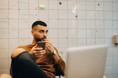 Young male programmer looking away while sitting with smart phone against wall at office