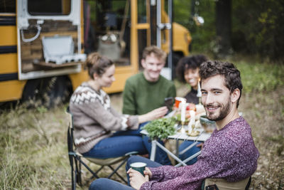 Portrait of smiling man sitting with friends at table during camping in forest