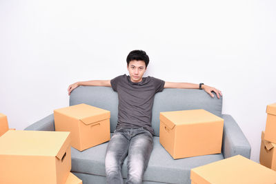 Portrait of young man with cardboard boxes sitting on sofa against wall at home