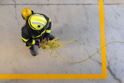 Top view of unrecognizable firefighter in protective hardhat and bright uniform standing on cement floor with rope during routine practices at work