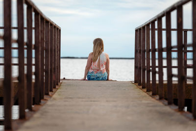 Rear view of woman sitting on pier over lake against sky