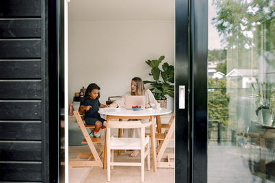 Working mother assisting son doing homework sitting at home seen through sliding door