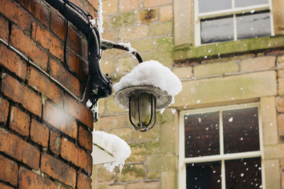 Low angle view of electric lamp mounted on brick wall during snowfall