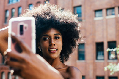 Young woman with afro hairstyle taking selfie through smart phone against building